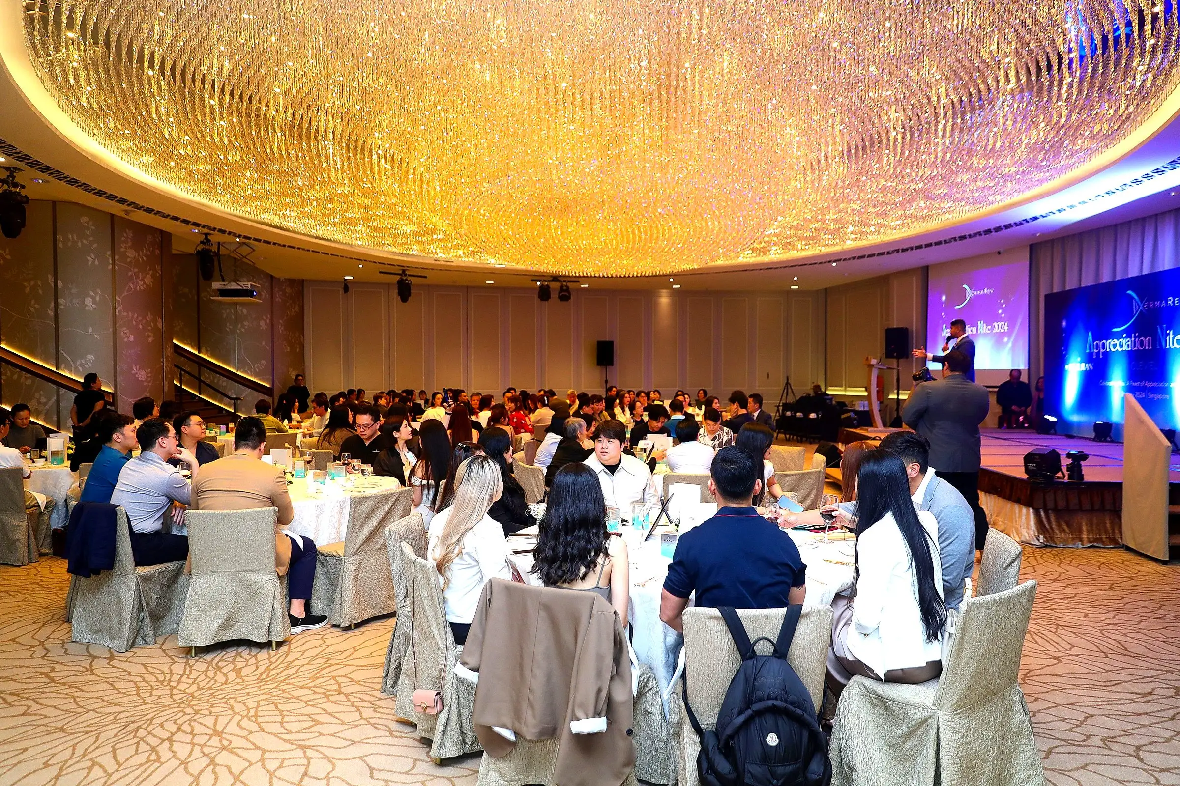 Guests seated in a hotel ballroom beneath a ring of chandeliers