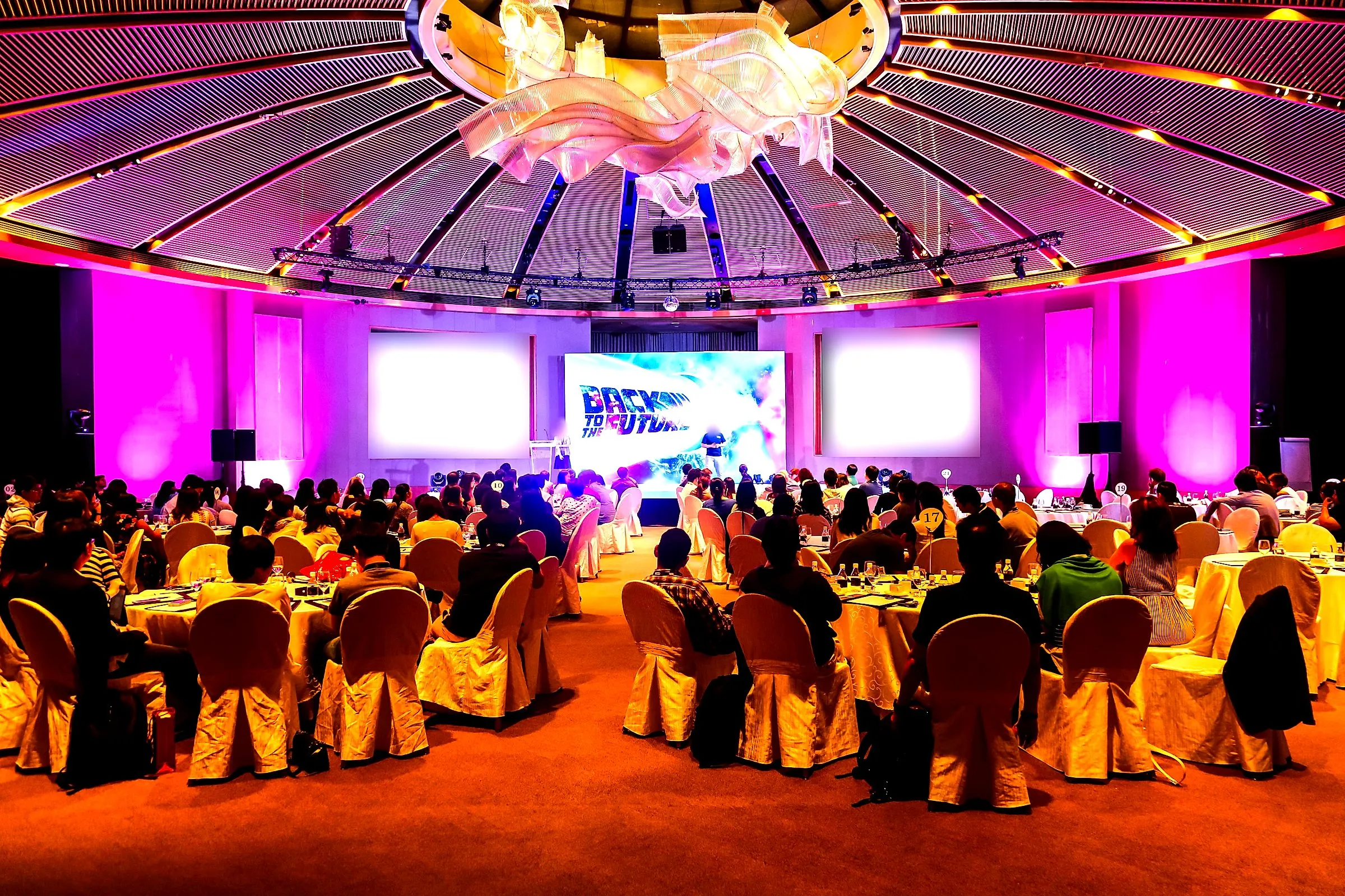 Seated dinner under a domed ceiling lit in magenta, with a themed stage screen