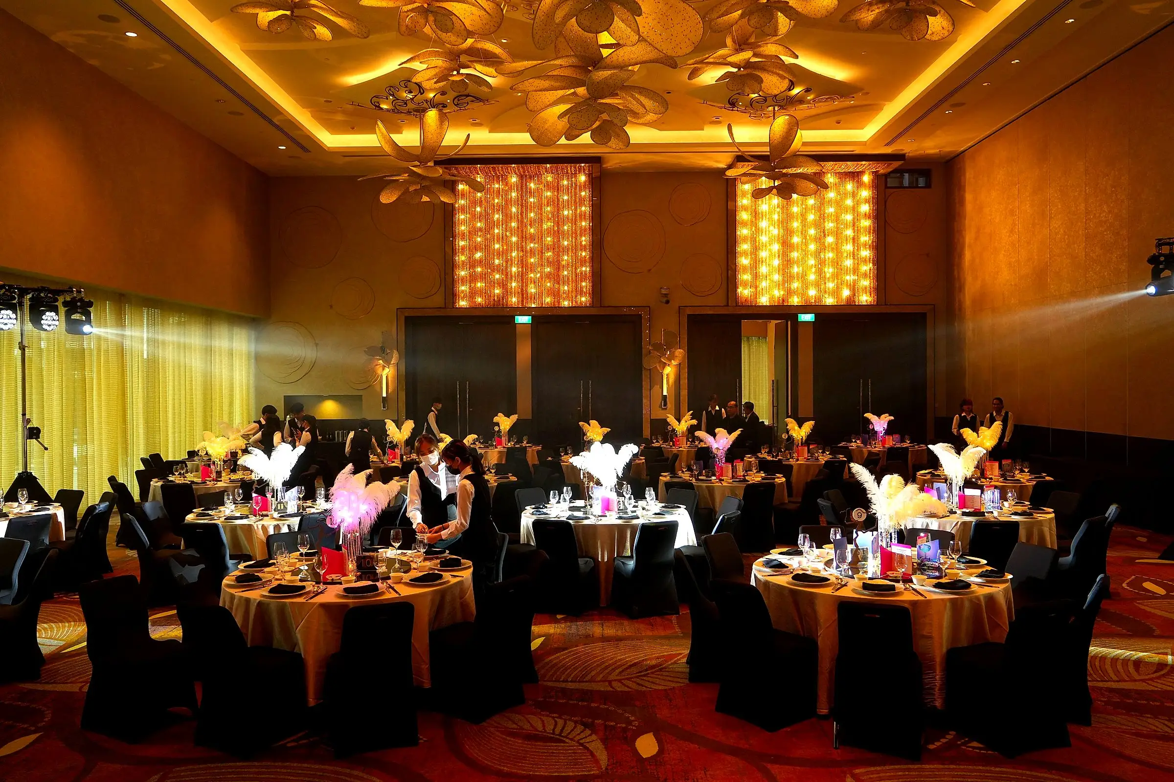 Wide view of a gala ballroom set with round tables, tall feather centrepieces and warm lighting