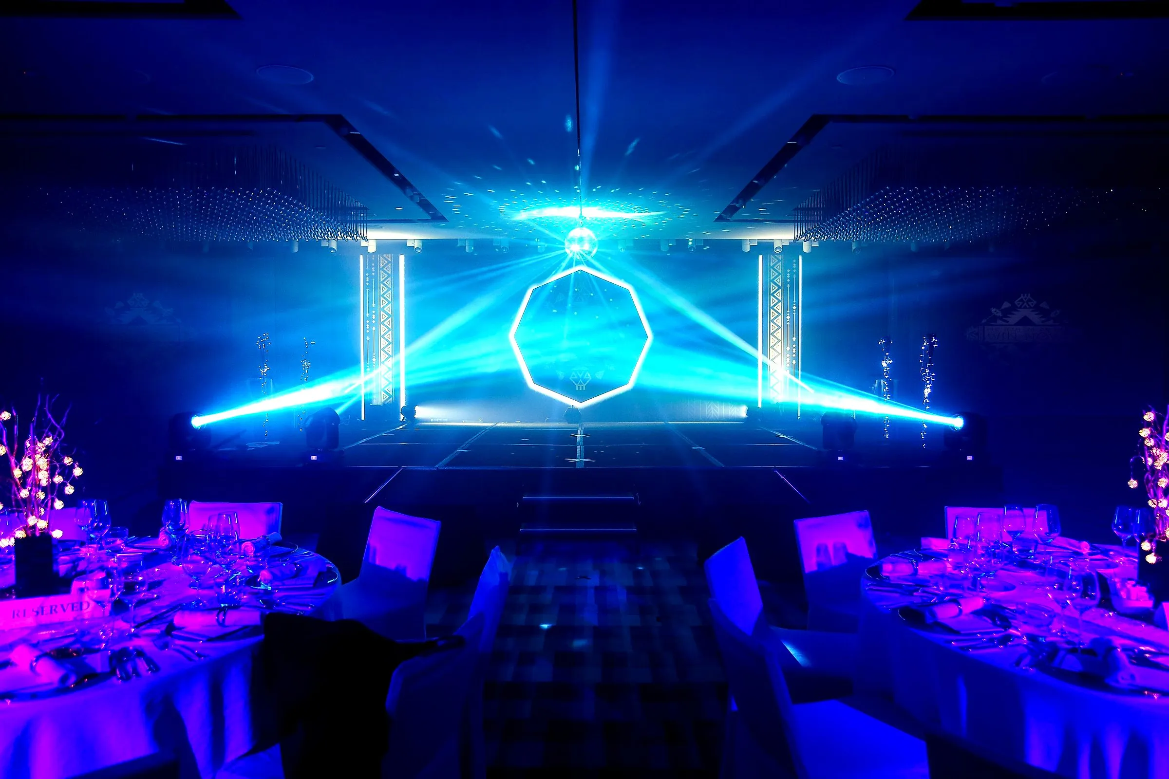Ballroom stage lit by blue laser beams and a mirror ball above dressed dinner tables